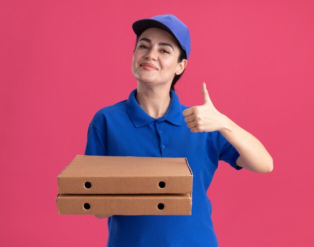 Zufriedene junge Lieferfrau in Uniform und Mütze mit Pizzapaketen, die Daumen nach oben zeigen