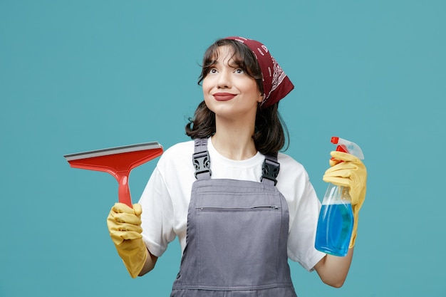 Zufriedene junge Putzfrau mit einheitlichem Bandana und Gummihandschuhen, die Wischer und Reinigungsmittel halten und isoliert auf blauem Hintergrund nach oben schauen