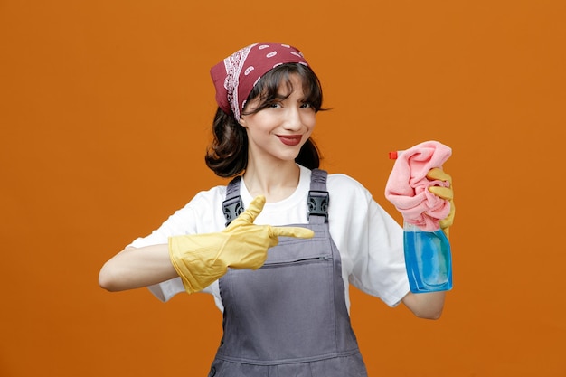 Zufriedene junge Putzfrau mit einheitlichen Gummihandschuhen und Bandana mit Stoffstaubtuch und Reinigungsmittel, die auf sie zeigt und auf die Kamera isoliert auf orangefarbenem Hintergrund blickt