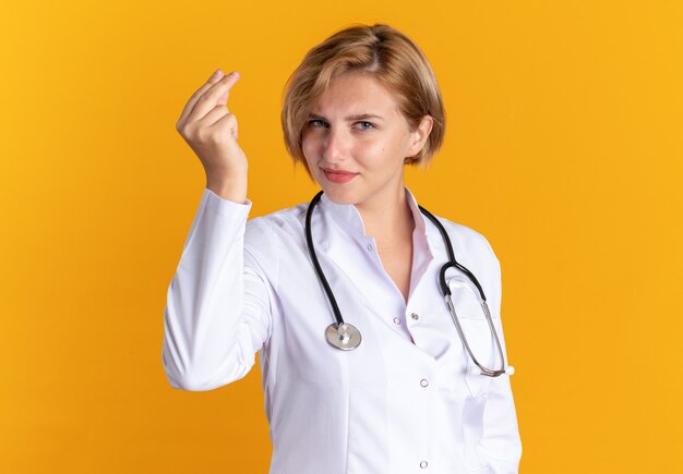 Zufriedene junge Ärztin, die ein medizinisches Gewand mit Stethoskop trägt, das die Spitzengeste isoliert auf der orangefarbenen Wand zeigt