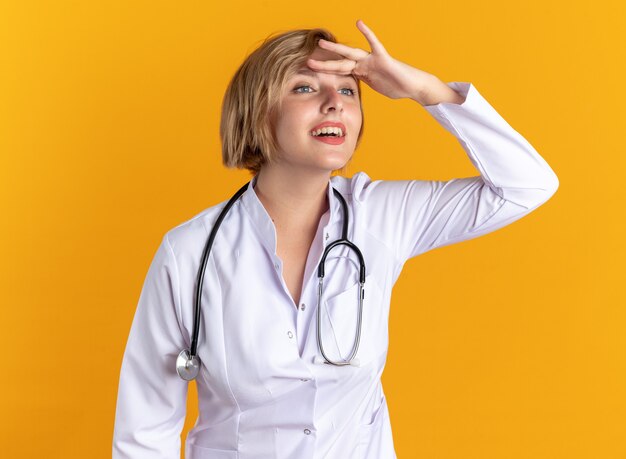 Zufriedene junge Ärztin, die medizinische Robe mit Stethoskop trägt und mit der Hand auf die orangefarbene Wand blickt