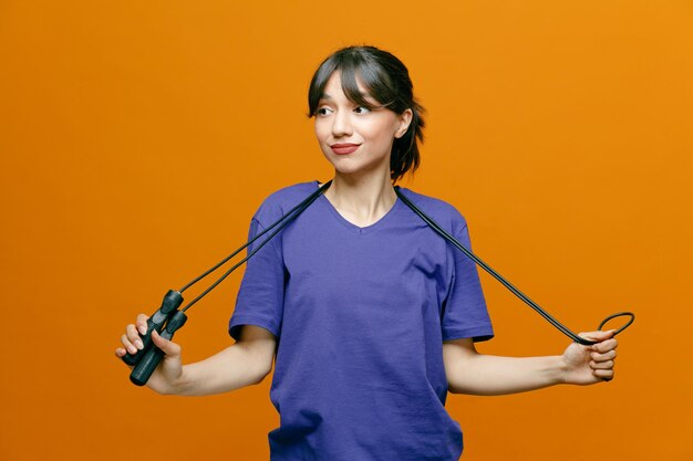 Zufriedene junge sportliche Frau mit T-Shirt, die Springseil hinter den Hals legt und die Seite isoliert auf orangefarbenem Hintergrund betrachtet