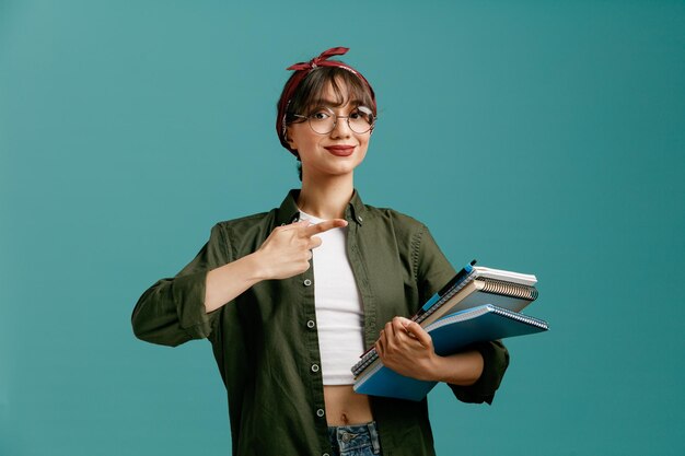 Zufriedene junge Studentin mit Bandana-Brille, die große Notizblöcke mit Stift hält und in die Kamera blickt, die auf Notizblöcke zeigt, die auf blauem Hintergrund isoliert sind