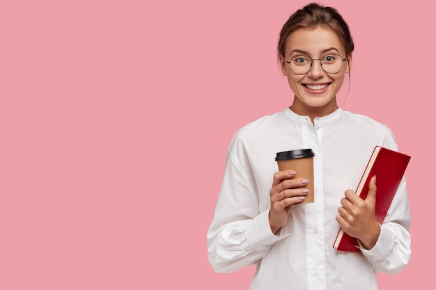 Zufriedene kaukasische Frau mit fröhlichem Ausdruck, trägt Kaffee zum Mitnehmen und Lehrbuch, hat positiven Ausdruck