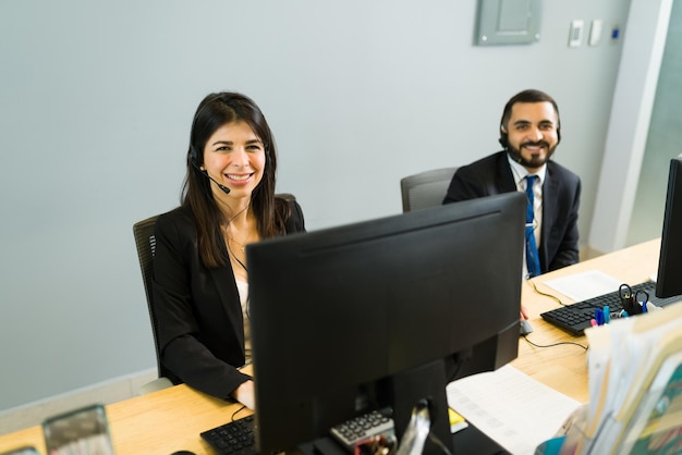 Zufriedene Kollegen, die lächeln und Augenkontakt herstellen, während sie an ihrem Schreibtisch in einem Callcenter sitzen. Führungskräfte warten auf Anrufe von Kunden für technischen Support