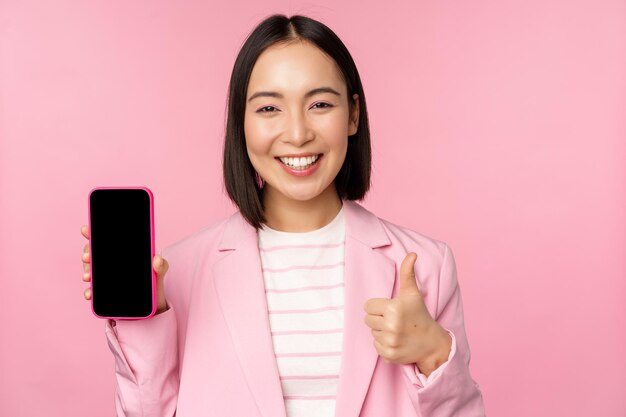 Zufriedene lächelnde asiatische Geschäftsfrau, die Handy-App-Website-Unternehmen auf dem Smartphone mit Bildschirm und Daumen nach oben rosa Hintergrund empfiehlt