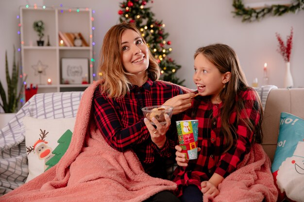 zufriedene Mutter füttert ihre Tochter mit Keksen, die auf der mit Decke bedeckten Couch sitzen und die Weihnachtszeit zu Hause genießen