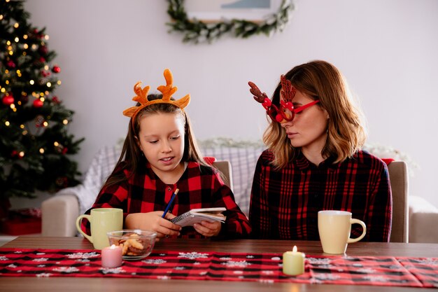 zufriedene Mutter in Rentierbrille sieht Tochter mit Bleistift und Notizbuch am Tisch sitzen und genießt die Weihnachtszeit zu Hause