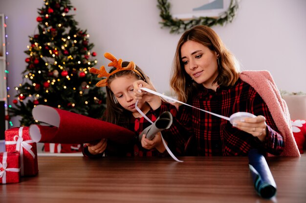 zufriedene mutter und tochter verpacken geschenke in bunte papiere zusammen sitzen am tisch und genießen die weihnachtszeit zu hause