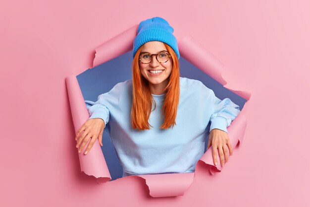 Zufriedene rothaarige europäische Frau lächelt angenehm, hat weiße Zähne und sommersprossige Haut trägt eine Brille mit blauem Hut und Sweatshirtständer in einem zerrissenen Loch aus rosa Papier