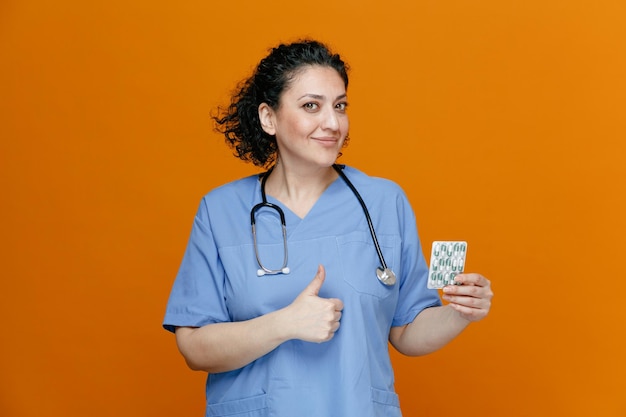 zufriedene Ärztin mittleren Alters, die Uniform und Stethoskop um den Hals trägt und eine Packung Pillen und Daumen nach oben zeigt, die isoliert auf orangefarbenem Hintergrund in die Kamera blickt