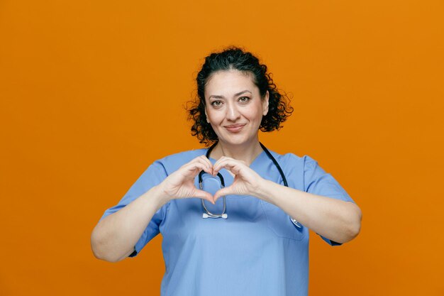 Zufriedene Ärztin mittleren Alters, die Uniform und Stethoskop um den Hals trägt und in die Kamera schaut, die ein Herzzeichen isoliert auf orangefarbenem Hintergrund zeigt