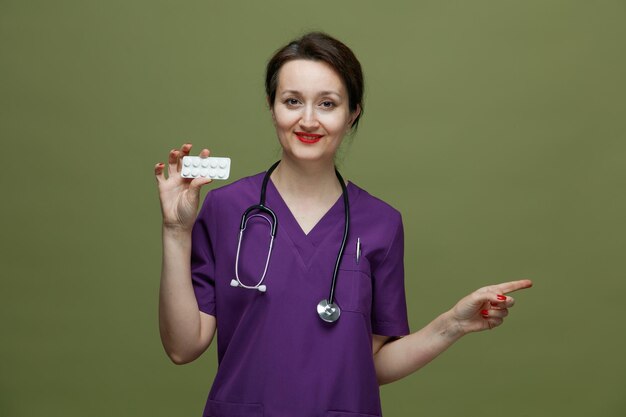 Zufriedene Ärztin mittleren Alters in Uniform und Stethoskop um den Hals, die in die Kamera blickt und eine Packung Tabletten zeigt, die auf olivgrünem Hintergrund isoliert zur Seite zeigen