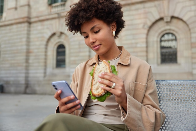 Zufriedene schöne lockige Frau isst Sandwich und benutzt das Handy zum Surfen im Internet oder zum Online-Chatten sitzt auf einer Bank gegen Stadtgebäude. Menschen essen Lifestyle und Technologiekonzept