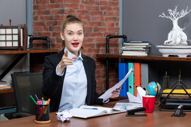Zufriedene und positive junge Dame, die an einem Tisch sitzt und das Dokument nach vorne im Büro hält