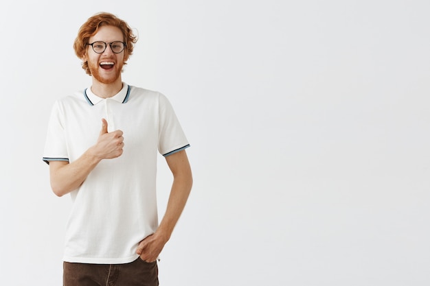 Zufriedener bärtiger Rotschopf, der mit Brille gegen die weiße Wand posiert