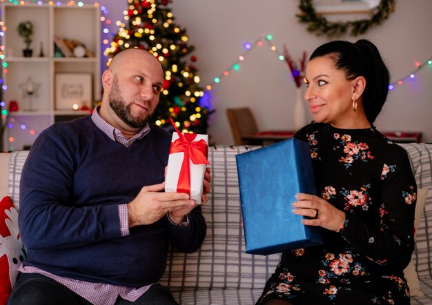 Zufriedener Ehemann und Ehefrau zu Hause in der Weihnachtszeit sitzen auf dem Sofa im Wohnzimmer, beide halten ein Geschenkpaket und schauen sich an
