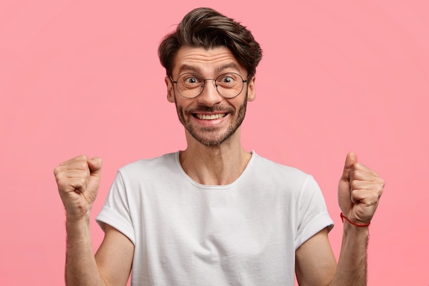 Zufriedener emotionaler unrasierter Mann mit trendigem Haarschnitt, dicken Stoppeln, geballten Fäusten mit Erfolg und Sieg, trägt weißes T-Shirt und Brille, isoliert über rosa Raum