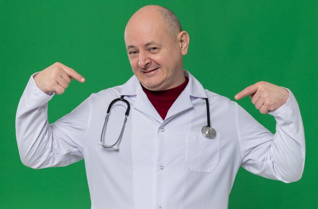 Zufriedener erwachsener Mann in Arztuniform mit Stethoskop, das auf sich selbst zeigt