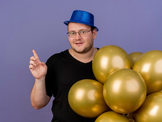 Zufriedener erwachsener slawischer Mann in optischer Brille mit blauem Partyhut steht mit Heliumballons nach oben