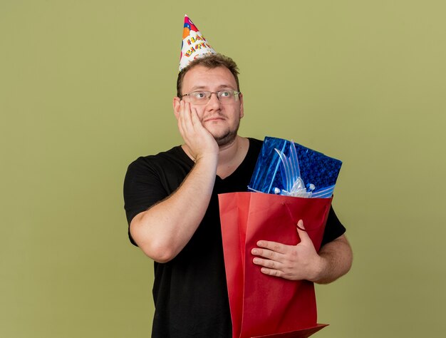 Zufriedener erwachsener slawischer Mann in optischer Brille mit Geburtstagskappe legt die Hand auf das Gesicht und hält die Geschenkbox in einer Papiereinkaufstasche mit Blick auf die Seite