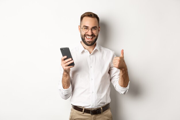 Zufriedener Geschäftsmann, der Daumen zeigt, nachdem er Handy benutzt hat, zufrieden stehend.