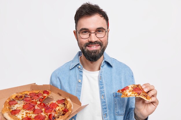 Zufriedener gutaussehender bärtiger Mann isst köstliche Pizza zum Abendessen fühlt sich hungrig, trägt runde Gläser und Hemd isst Junk Food