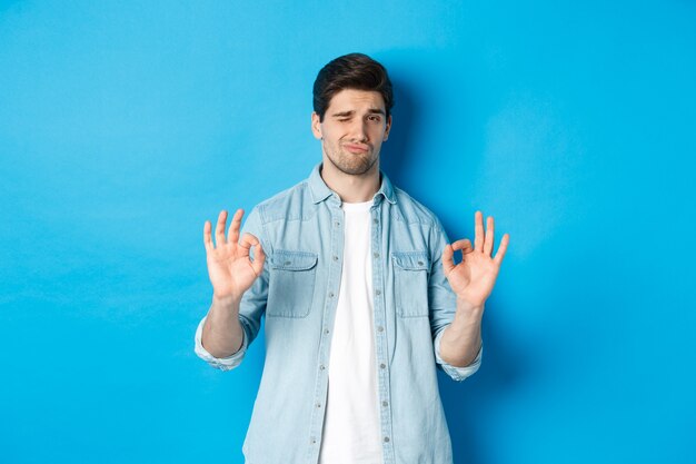 Zufriedener gutaussehender Mann, der A-OK-Zeichen zeigt und zufrieden aussieht, etwas Gutes genehmigt und vor blauem Hintergrund steht.