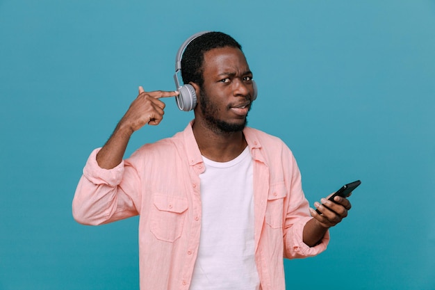 Zufriedener junger Afroamerikaner, der das Telefon mit Kopfhörern isoliert auf blauem Hintergrund hält