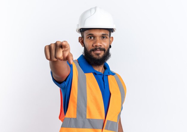 Zufriedener junger afroamerikanischer Baumeister in Uniform mit Schutzhelm, der auf die Kamera zeigt, die auf weißem Hintergrund mit Kopienraum isoliert ist