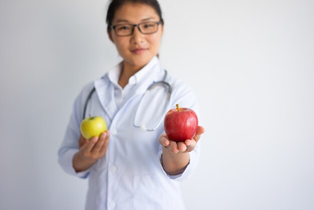 Zufriedener junger asiatischer weiblicher Doktor, der roten Apfel anbietet.