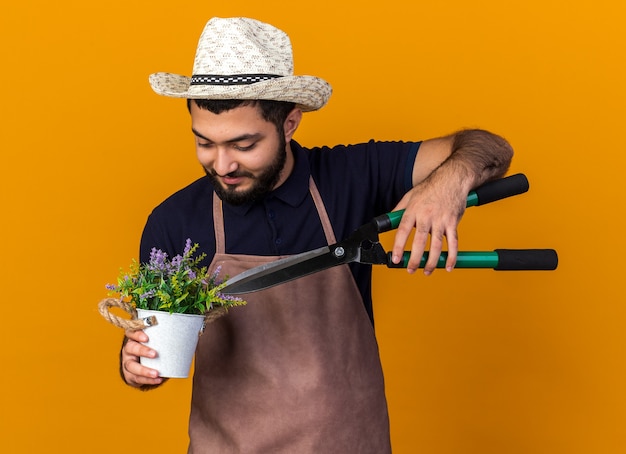 zufriedener junger kaukasischer männlicher Gärtner mit Gartenhut, der eine Gartenschere über Blumen im Blumentopf hält, isoliert auf oranger Wand mit Kopierraum