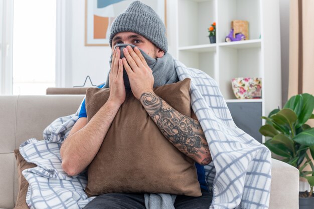 zufriedener junger kranker Mann mit Schal und Wintermütze, der in eine Decke gehüllt ist und auf dem Sofa im Wohnzimmer sitzt und ein Kissen hält, das den Mund mit einem Schal bedeckt