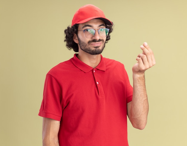 Zufriedener junger Liefermann in roter Uniform und Mütze mit Brille, der nach vorne schaut und Geldgeste einzeln auf olivgrüner Wand macht