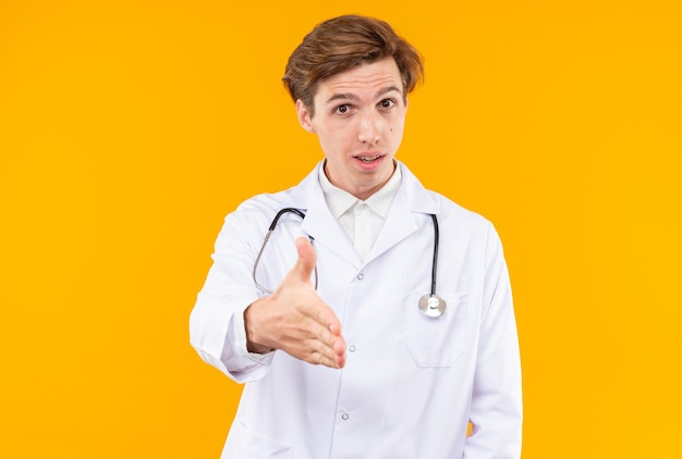 Zufriedener junger männlicher Arzt, der ein medizinisches Gewand mit Stethoskop trägt, das die Hand vor der Kamera hält, isoliert auf der orangefarbenen Wand?