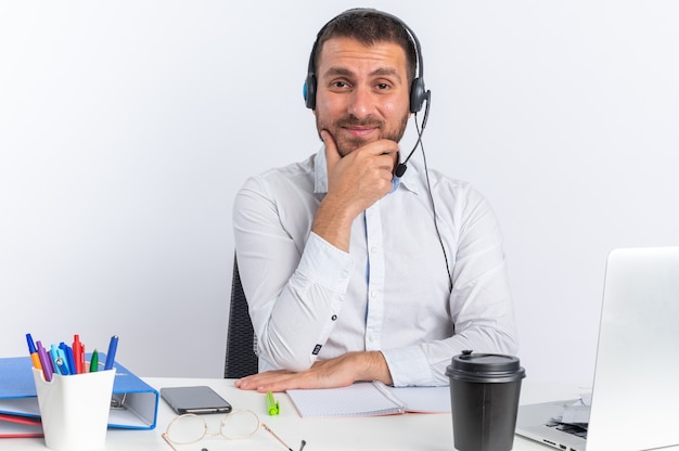 Zufriedener junger männlicher Callcenter-Betreiber mit Headset am Tisch sitzend mit Bürowerkzeugen packte das Kinn