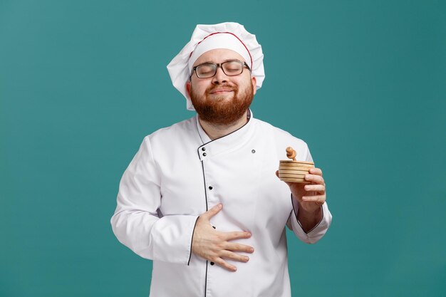 Zufriedener junger männlicher Koch mit einheitlicher Brille und Mütze, der eine Holzschale mit Honigschöpflöffel darin hält und die Hand auf dem Bauch hält, mit geschlossenen Augen isoliert auf blauem Hintergrund