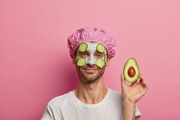 Zufriedener junger Mann hat saubere, frische Haut, hält ein Stück Avocado, verwendet frisches Gemüse für die Herstellung der Gesichtsmaske und trägt eine Duschhaube