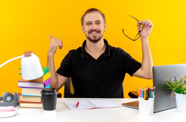 Zufriedener junger Student, der am Tisch mit Schulwerkzeugen sitzt und eine Brille hält, die den Daumen isoliert auf der orangefarbenen Wand zeigt