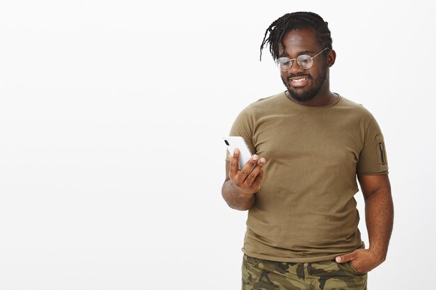 Zufriedener Kerl mit Brille, die mit seinem Telefon gegen die weiße Wand posiert