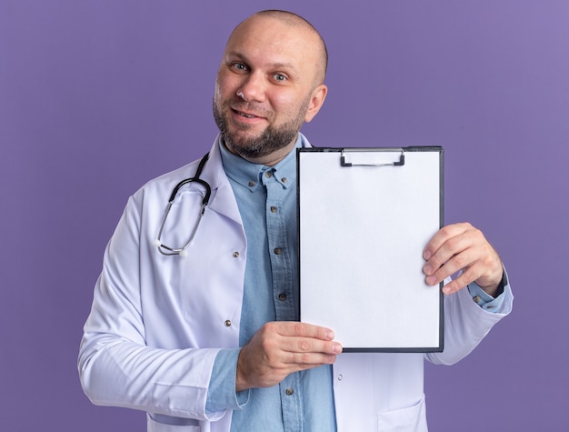Zufriedener männlicher Arzt mittleren Alters, der ein medizinisches Gewand und ein Stethoskop trägt, das die Zwischenablage nach vorne zeigt, die auf der violetten Wand isoliert ist?