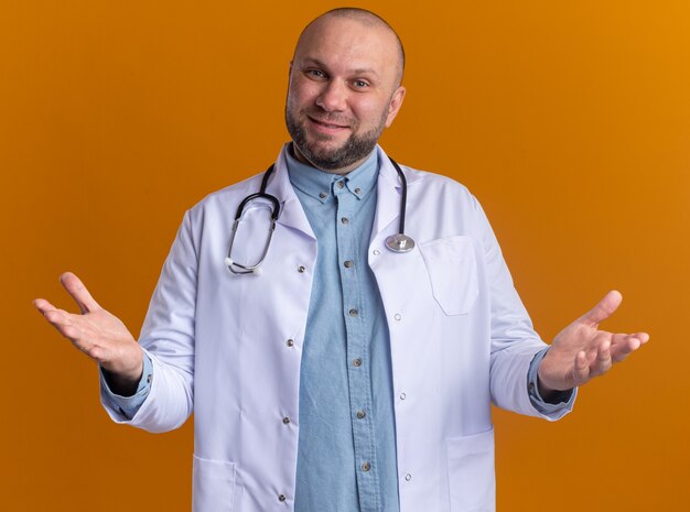 Zufriedener männlicher Arzt mittleren Alters, der ein medizinisches Gewand und ein Stethoskop trägt, das leere Hände zeigt