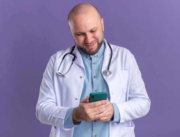 Zufriedener männlicher Arzt mittleren Alters, der ein medizinisches Gewand und ein Stethoskop trägt und sein Mobiltelefon isoliert auf der lila Wand benutzt