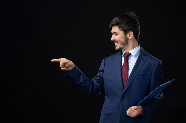 Zufriedener männlicher Büroangestellter im Anzug, der Dokumente hält und etwas auf der rechten Seite auf isolierte dunkle Wand zeigt