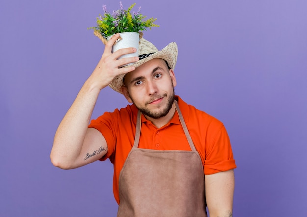 Kostenloses Foto zufriedener männlicher gärtner mit gartenhut hält blumentopf über dem kopf