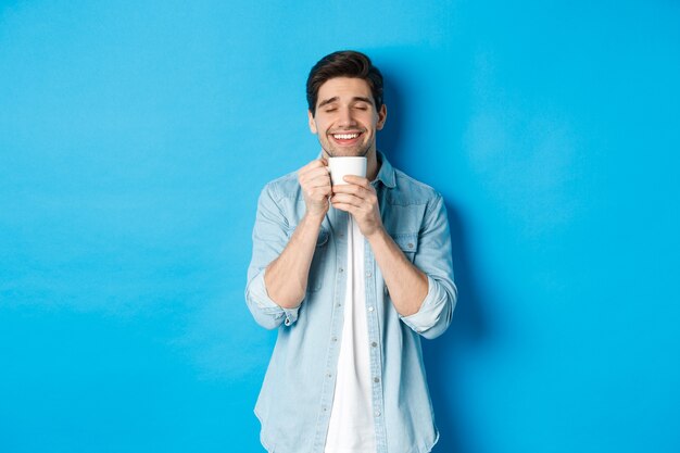 Zufriedener Mann, der eine Tasse Tee oder Kaffee genießt, eine Tasse mit einem zufriedenen Lächeln hält und vor blauem Hintergrund steht