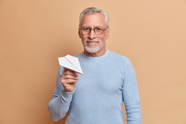 Zufriedener, selbstbewusster, reifer, grauhaariger Mann mit einem Lächeln hält handgeschöpftes Papierflugzeug, das sicher ist, dass es in erfolgreicher Zukunft eine Brille und einen blauen Pullover trägt, die über der braunen Wand isoliert sind