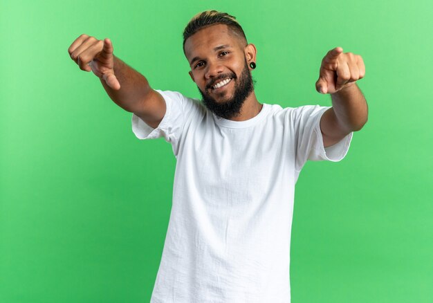 Zufriedener und glücklicher afroamerikanischer junger Mann im weißen T-Shirt mit Blick auf die Kamera, die mit den Zeigefingern auf die Kamera zeigt