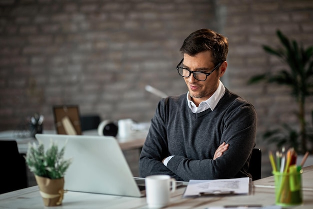 Zufriedener Unternehmer mit verschränkten Armen, der während der Arbeit im Büro gute Nachrichten auf einem Computer liest