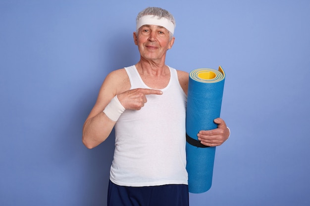 Zufriedener weißhaariger mann mit yogamatte, die isoliert posiert, mit zeigefinger beiseite zeigt und ärmelloses t-shirt, haarband und armband trägt.